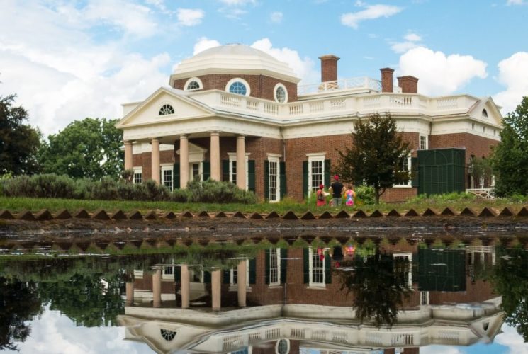 Historic brick home with cream trim, several windows and columns in front of large pond