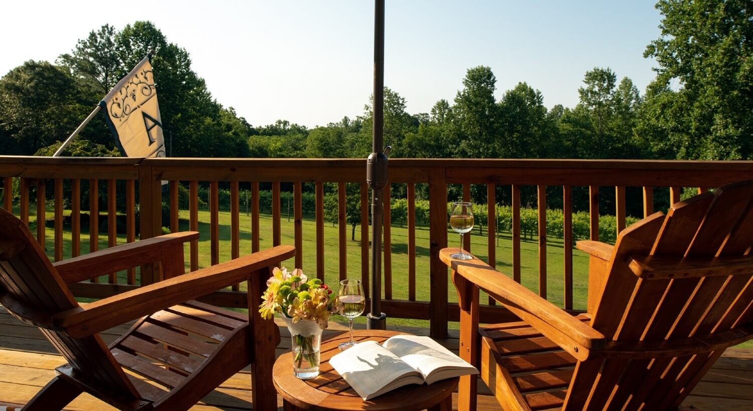 Outdoor deck with two adirondack chairs, table with book and wine glass overlooking a lawn and trees