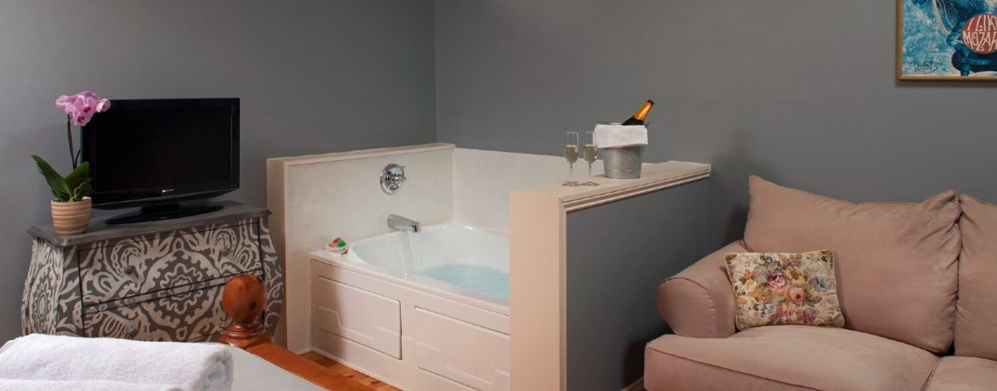 Corner jacuzzi tub in room with tan loveseat and dresser with TV