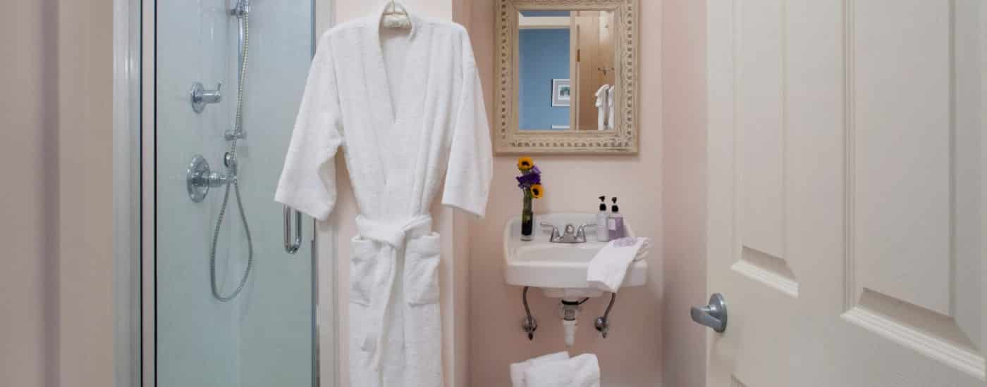 Bathroom with single white sink under framed mirror, hanging white robe and stand up shower