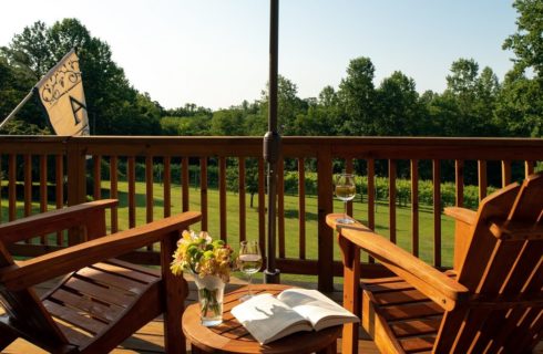 Outdoor deck with two adirondack chairs, table with book and wine glass overlooking a lawn and trees