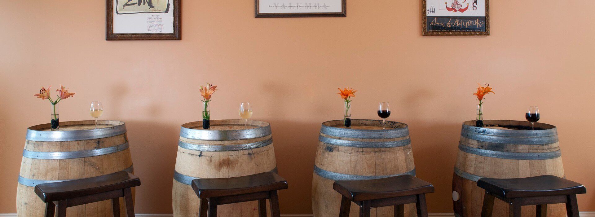 Four large wooden barrels with metal trim each with a vase of flowers and glass of wine