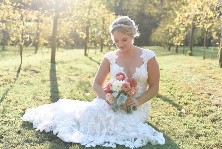 Bride in white gown, holding a bouquet and sitting on grass with sunlight coming through vineyard trees