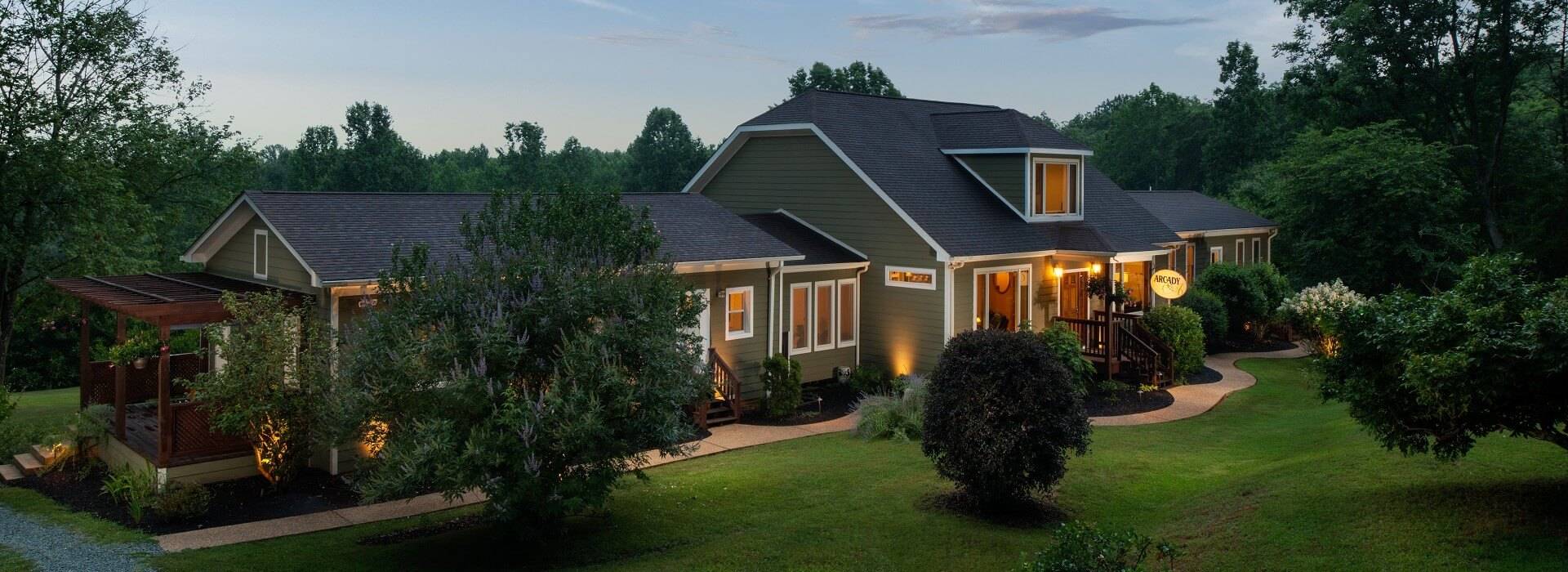 Outside of an expansive elegant home with green siding, warm lighting and large grassy lawn with trees